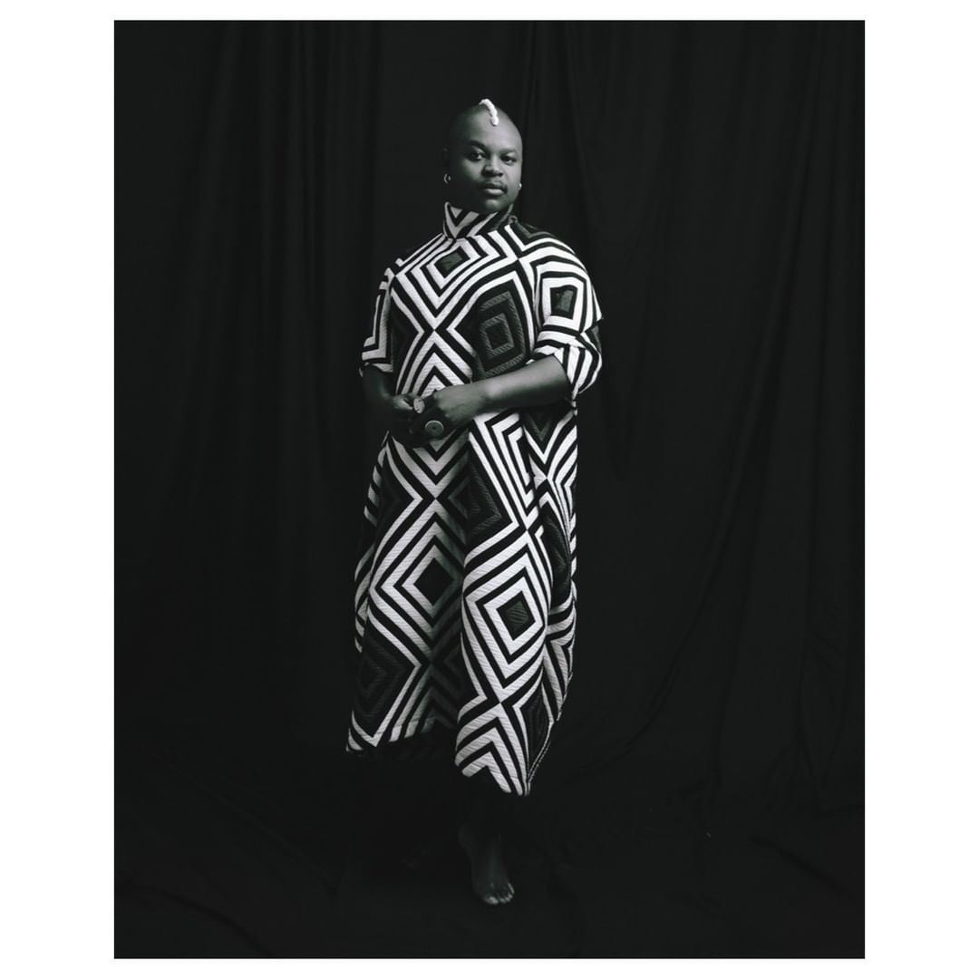 Studio portrait of adult Black woman wearing patterned tunic, standing against a black curtain backdrop in black and white.