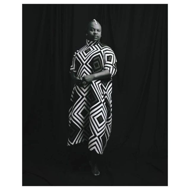Studio portrait of adult Black woman wearing patterned tunic, standing against a black curtain backdrop in black and white.