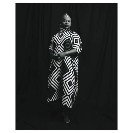 Studio portrait of adult Black woman wearing patterned tunic, standing against a black curtain backdrop in black and white.