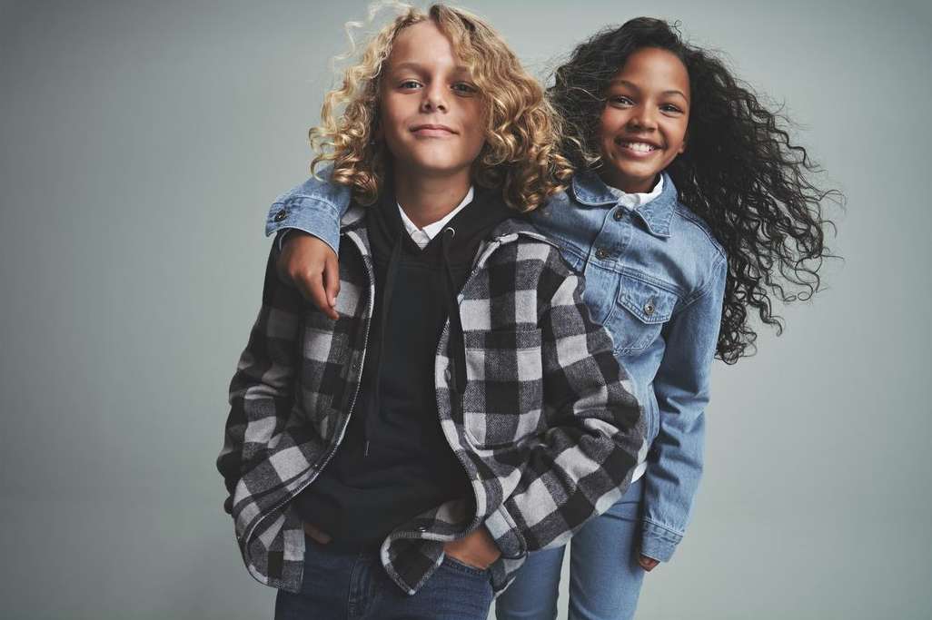 Two smiling children, a boy and a girl, pose indoors in denim jackets and plaid shirt against a neutral gray backdrop.