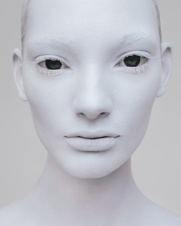Close-up portrait of a female with face and body painted white, contrasting with her green eyes, against a light gray background.