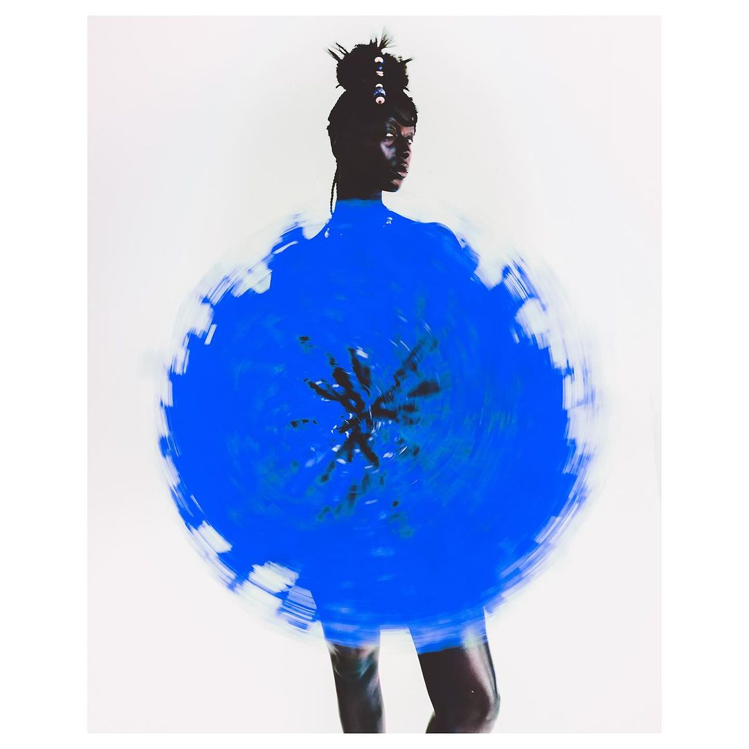 Artistic studio shot of a woman with dark skin wearing a blue circular garment against a neutral background. Long exposure effect.