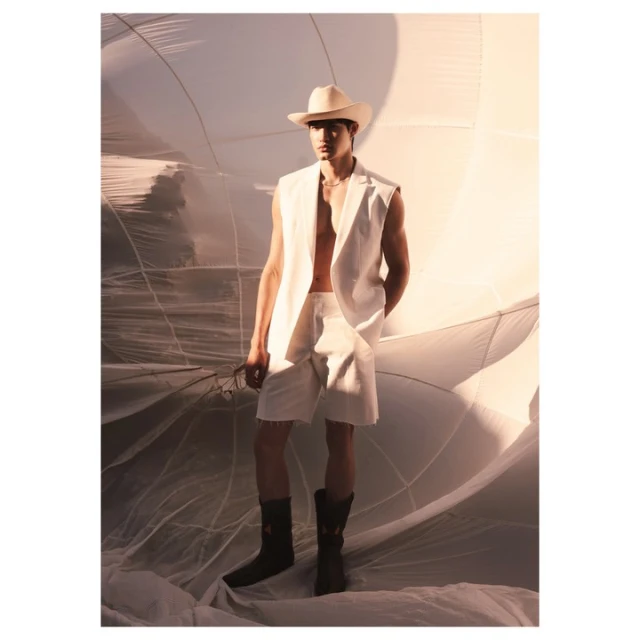 A man in a white sleeveless suit and cowboy hat poses against a wrinkled white backdrop, wearing black cowboy boots in a fashion editorial.