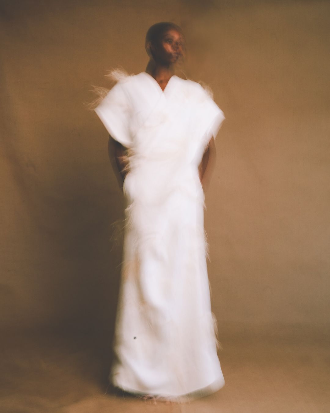 Model wearing a floor-length white textured gown with feathered edges against a warm neutral studio backdrop.