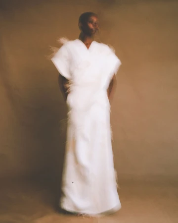 Model wearing a floor-length white textured gown with feathered edges against a warm neutral studio backdrop.