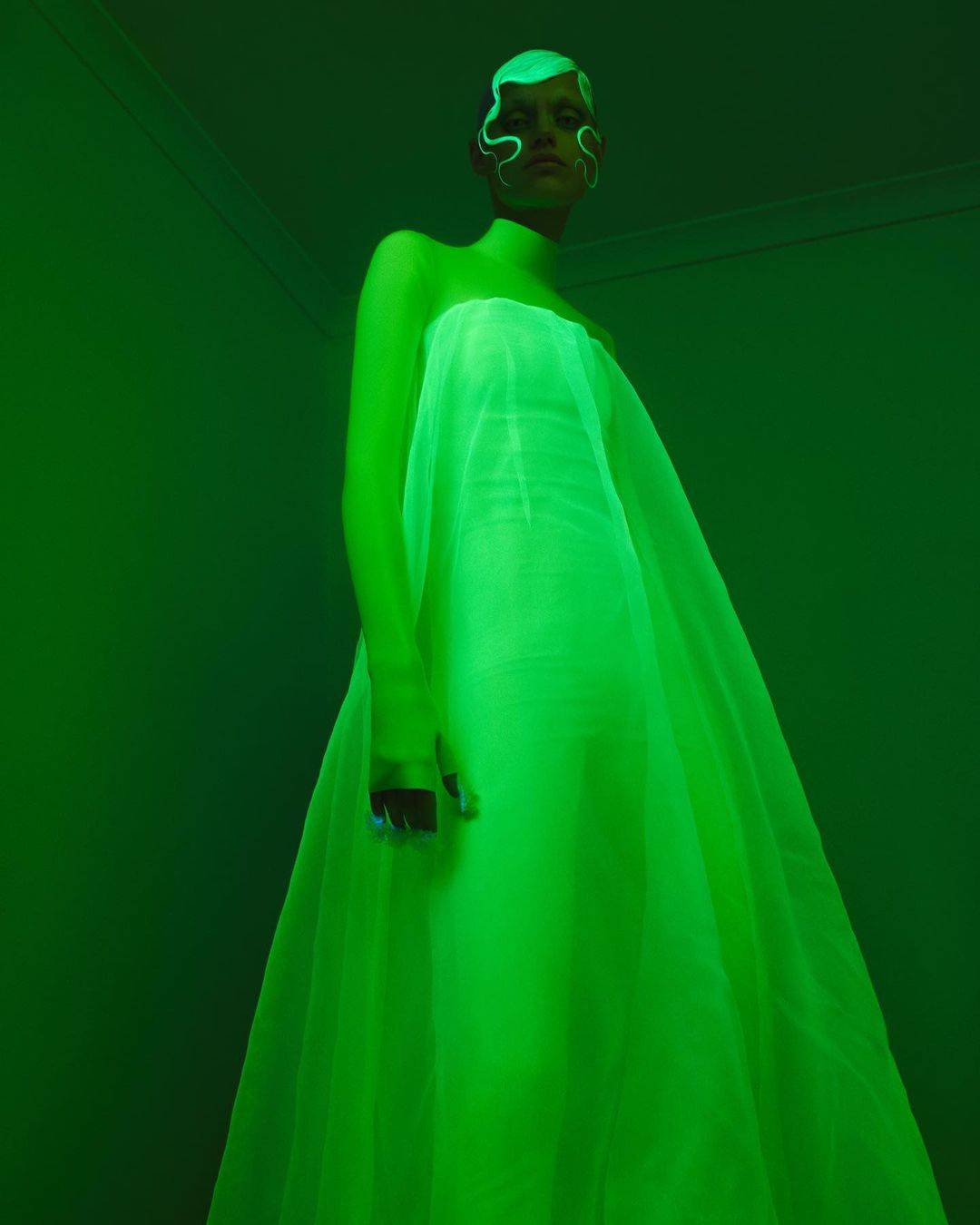 Woman in a long, light-green gown, standing in a room with a green glow. Her hair and face are also highlighted in green.