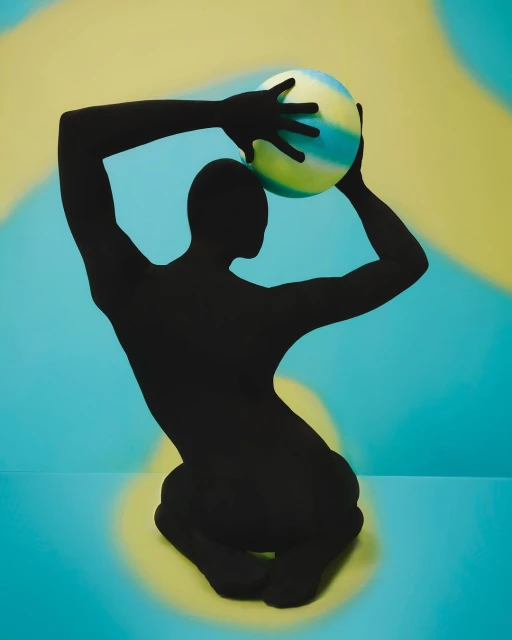 Black-clad figure kneels while holding a green, white, and blue ball, set against a yellow and blue mottled backdrop in a studio shot.