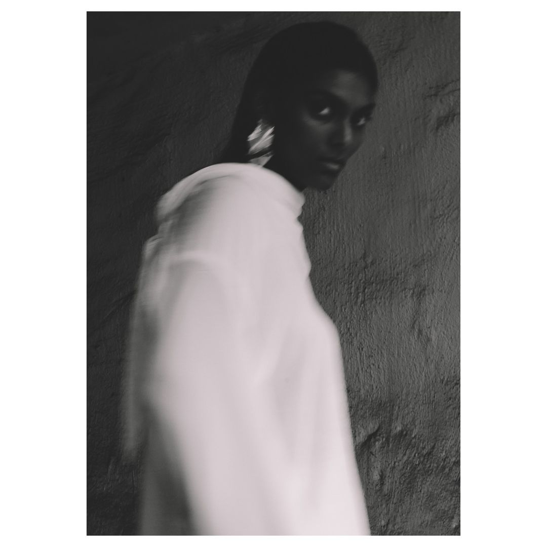 Monochromatic portrait of an adult woman in a blurred white top, with dark hair and statement earrings, against a textured gray wall.