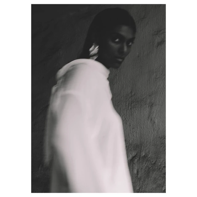 Monochromatic portrait of an adult woman in a blurred white top, with dark hair and statement earrings, against a textured gray wall.
