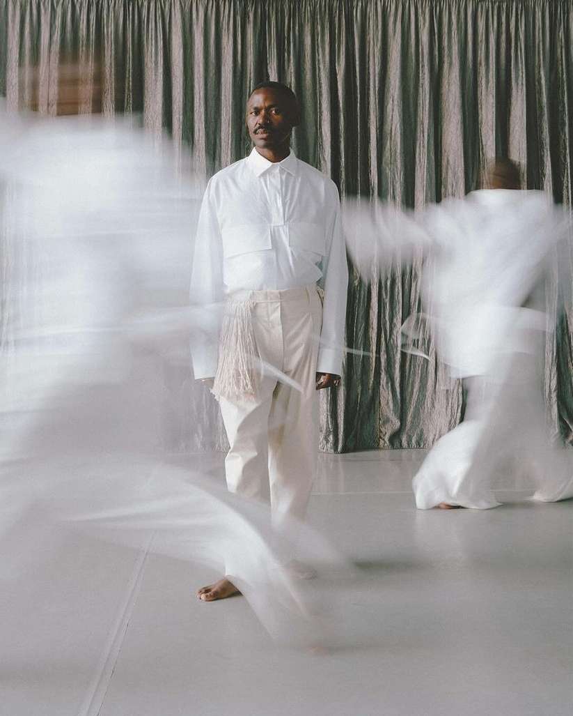 Man in white shirt and pants stands on a white floor in front of curtain with blurred figures in white clothing in motion. 