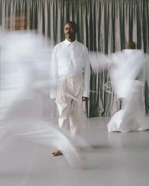 Man in white shirt and pants stands on a white floor in front of curtain with blurred figures in white clothing in motion. 