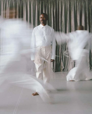 Man in white shirt and pants stands on a white floor in front of curtain with blurred figures in white clothing in motion. 
