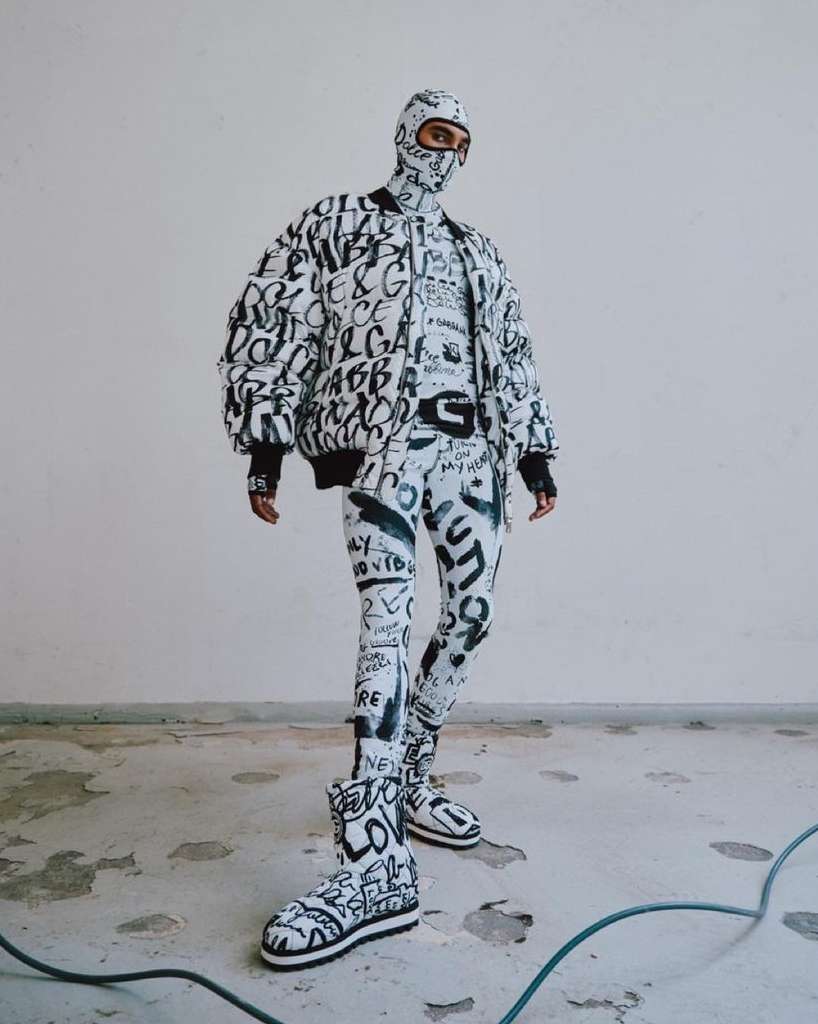 Person in full Dolce & Gabbana patterned outfit with jacket, balaclava, leggings, and boots, posing in a white indoor setting.