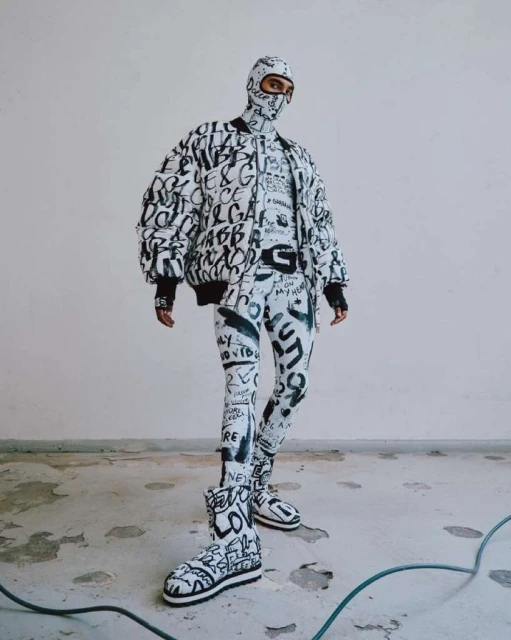 Person in full Dolce & Gabbana patterned outfit with jacket, balaclava, leggings, and boots, posing in a white indoor setting.