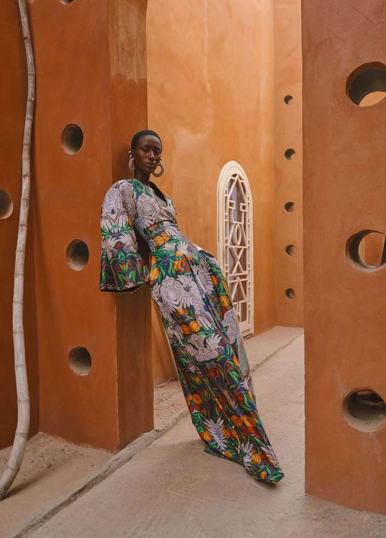 Elegant Black woman in floral maxi dress leans against orange wall with circular openings in a building with unique architectural details.