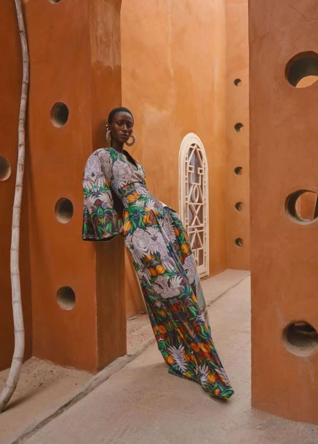 Elegant Black woman in floral maxi dress leans against orange wall with circular openings in a building with unique architectural details.