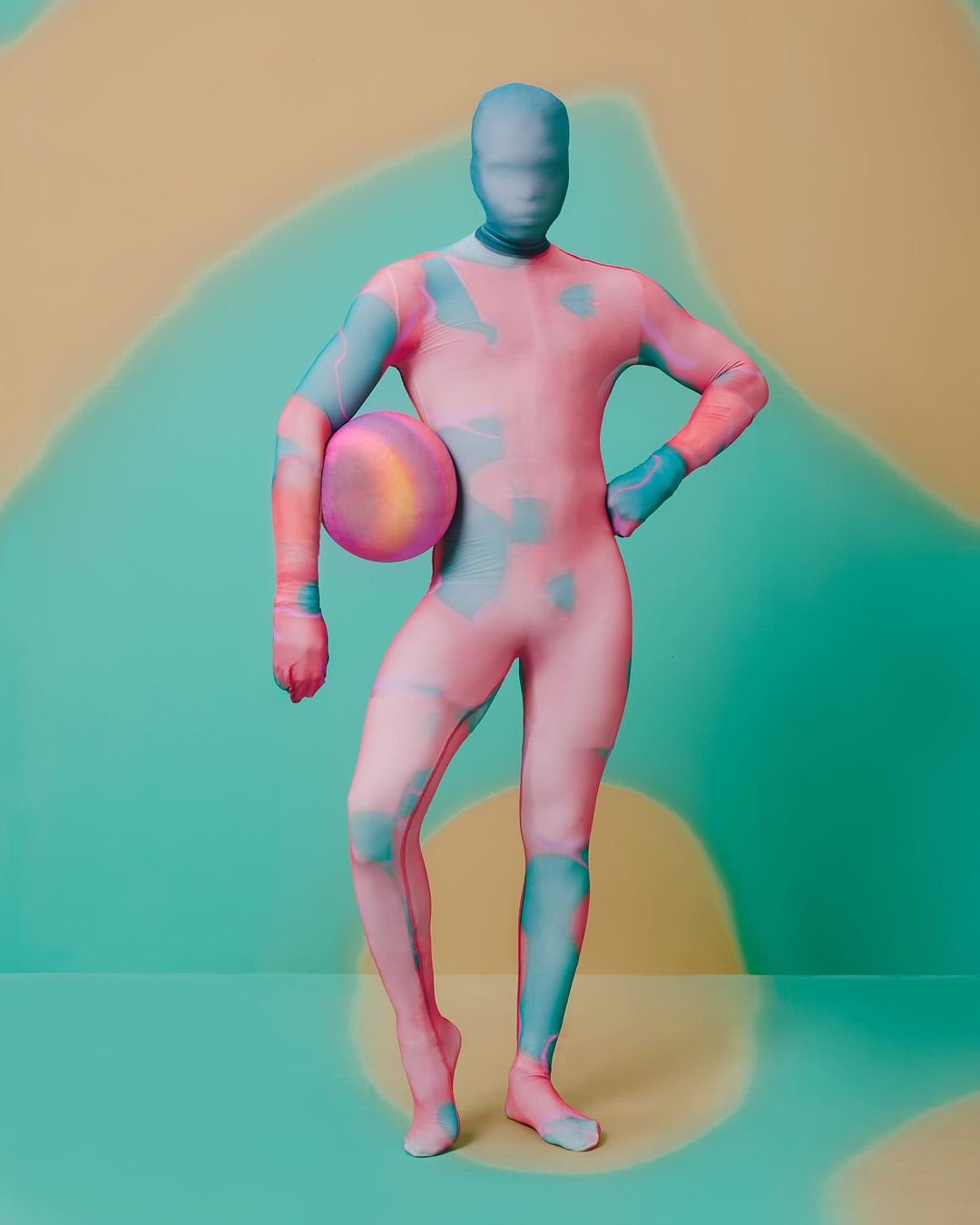 Full body studio shot of a person in a pink and blue morphsuit holding a ball in front of a patterned teal and tan backdrop.