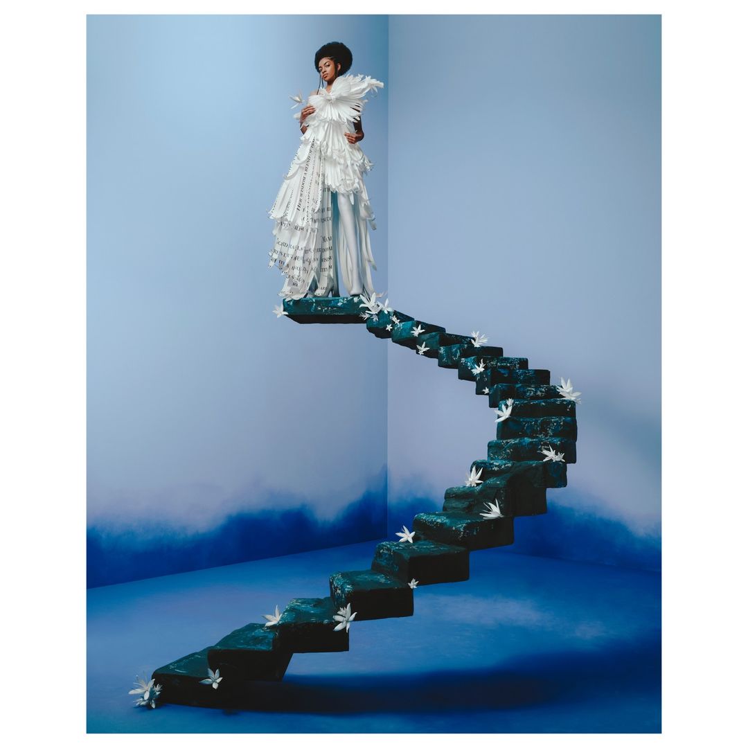 Fashionable woman in an ornate white feathered gown standing on a winding staircase against a studio backdrop with blue gradient.