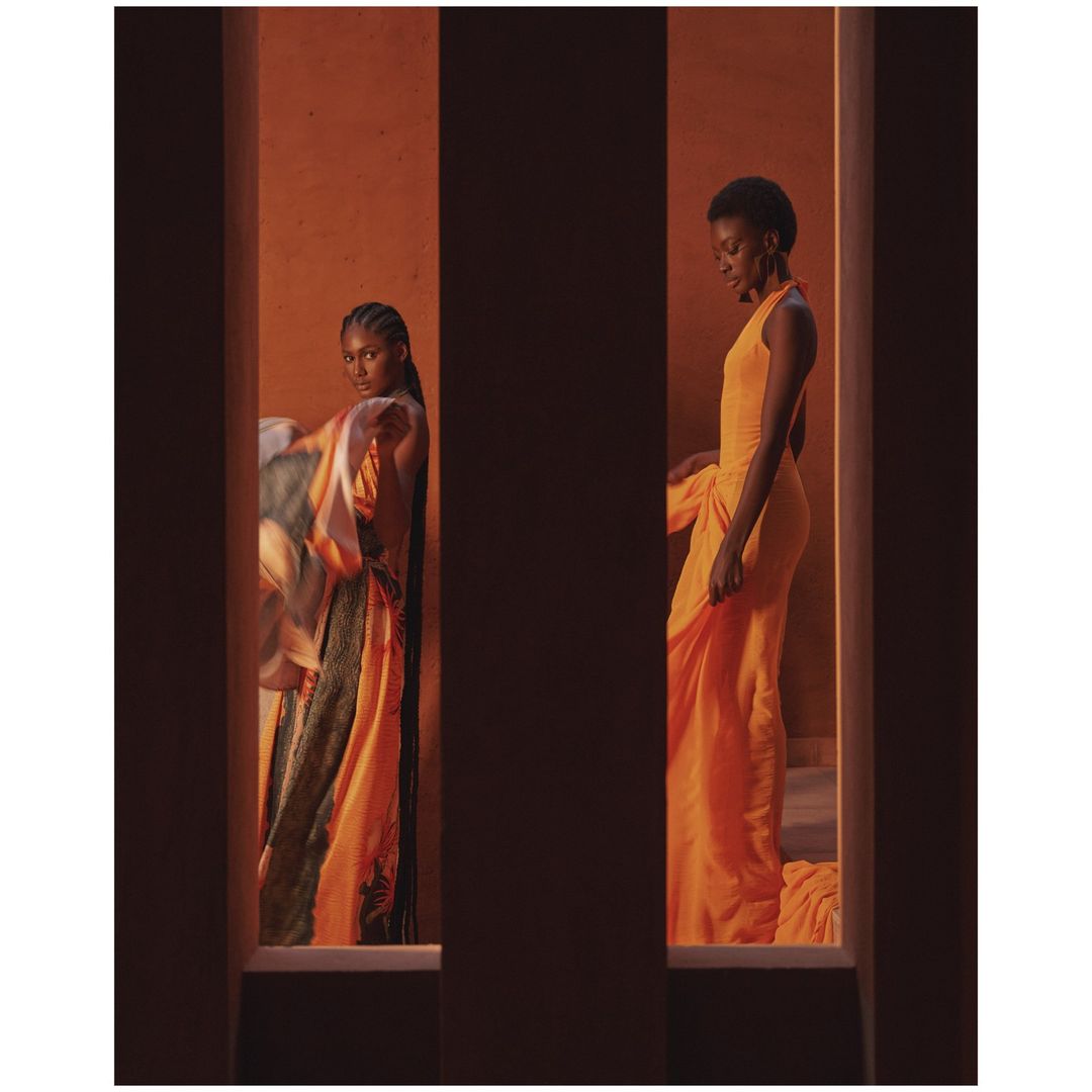 Two Black women in orange dresses strike poses framed by vertical architectural features, highlighting fashion and studio aesthetics.