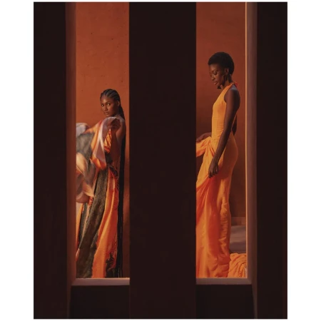 Two Black women in orange dresses strike poses framed by vertical architectural features, highlighting fashion and studio aesthetics.
