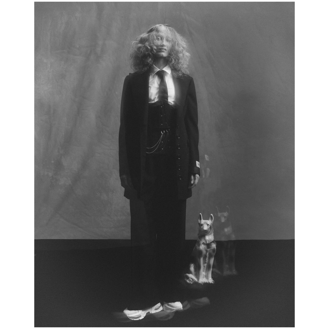 A person in a suit stands with a dog statue in a high-contrast black and white indoor studio portrait on a dark floor.