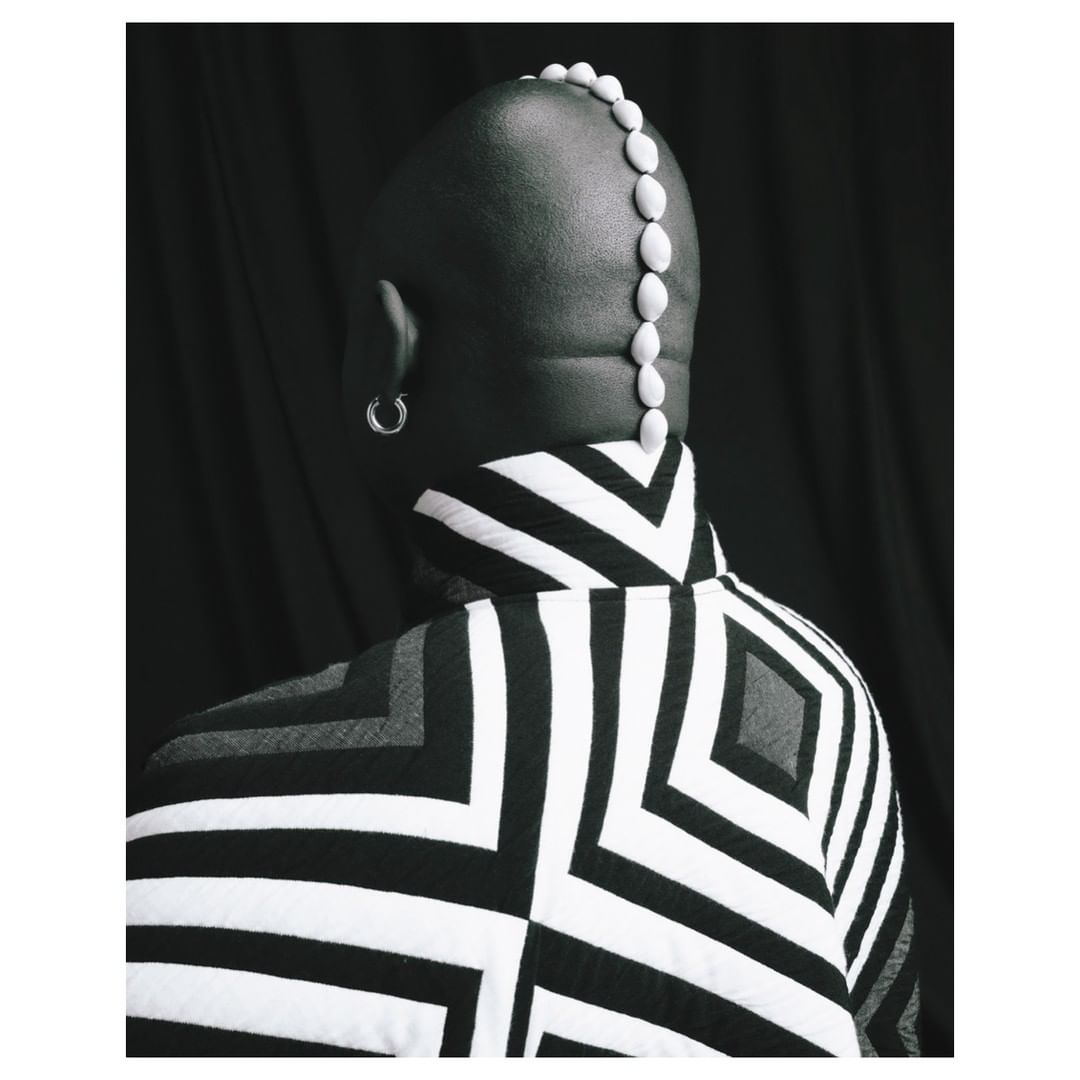 Back view of a bald person in a black-and-white patterned jacket with a row of white pebbles on their head against a dark backdrop.