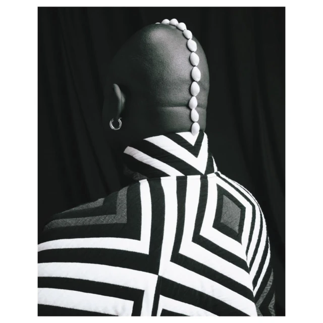 Back view of a bald person in a black-and-white patterned jacket with a row of white pebbles on their head against a dark backdrop.