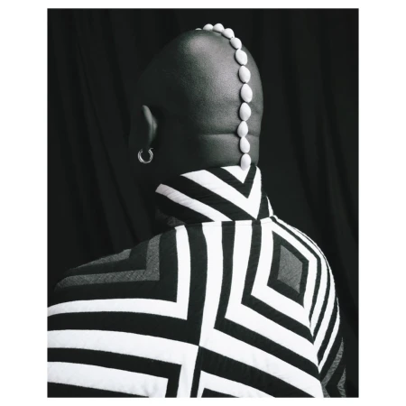 Back view of a bald person in a black-and-white patterned jacket with a row of white pebbles on their head against a dark backdrop.