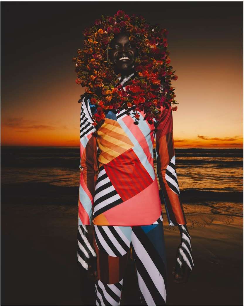 A cheerful Black model on a beach at sunset, adorned with colorful flowers, wearing a vibrant striped and patterned suit, radiating joy.