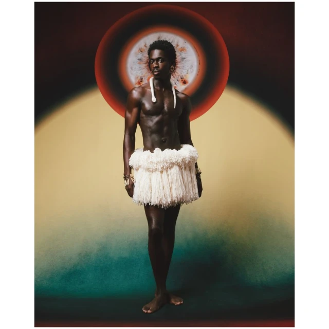 Striking studio shot of a Black male model in traditional wear: bare-chested, grass skirt, and beaded jewelry, against a vibrant backdrop.