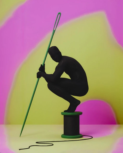 Surreal studio shot: a silhouetted figure in a crouching pose, perched on a thread spool, holds a giant green needle against a pink and yellow backdrop.