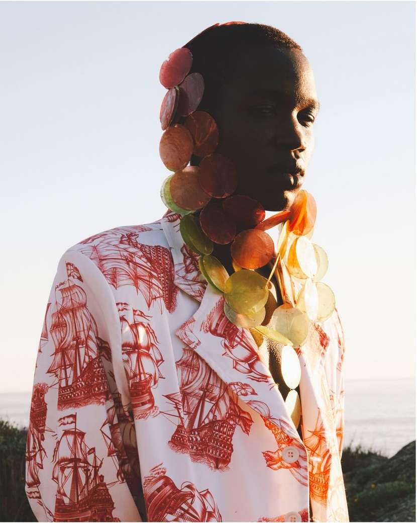 Eye-level shot of a Black male model in a ship-print blazer and round disc accessories posing outside against a sky and coastal backdrop.