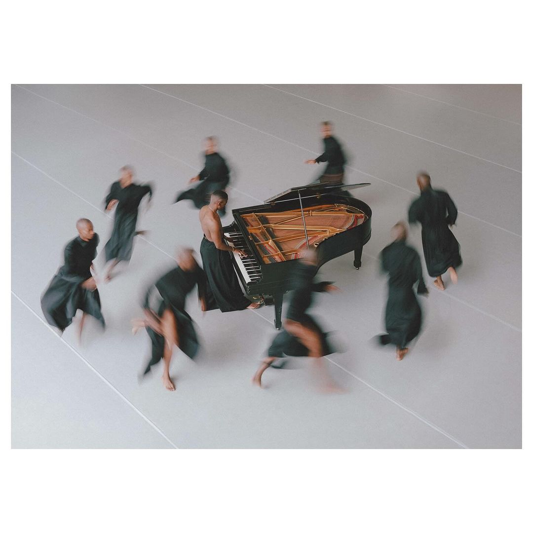 A grand piano takes center stage surrounded by multiple blurred figures in flowing black garments on a grey background in a studio setting.