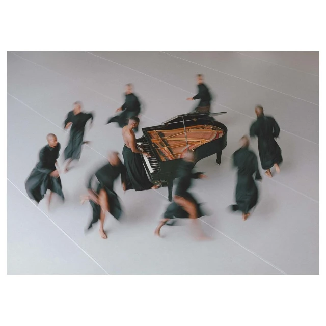 A grand piano takes center stage surrounded by multiple blurred figures in flowing black garments on a grey background in a studio setting.