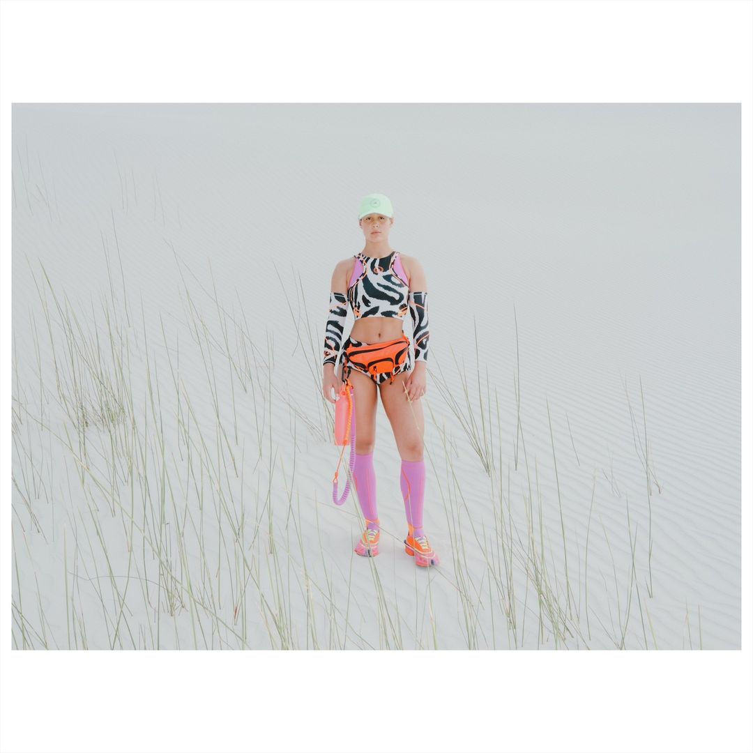 Adult female in a zebra print outfit with an orange fanny pack poses in a sandy outdoor setting with sparse vegetation.