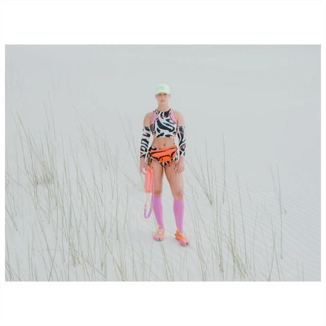 Adult female in a zebra print outfit with an orange fanny pack poses in a sandy outdoor setting with sparse vegetation.