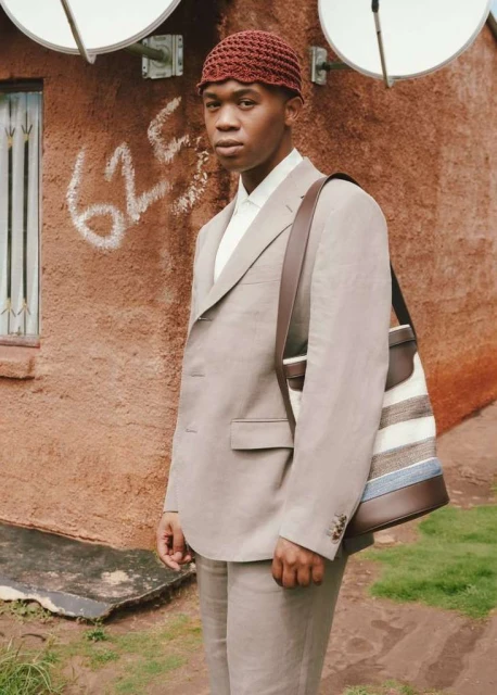 Man in a light-brown suit standing outdoors near a stucco wall with graffiti and two satellite dishes.