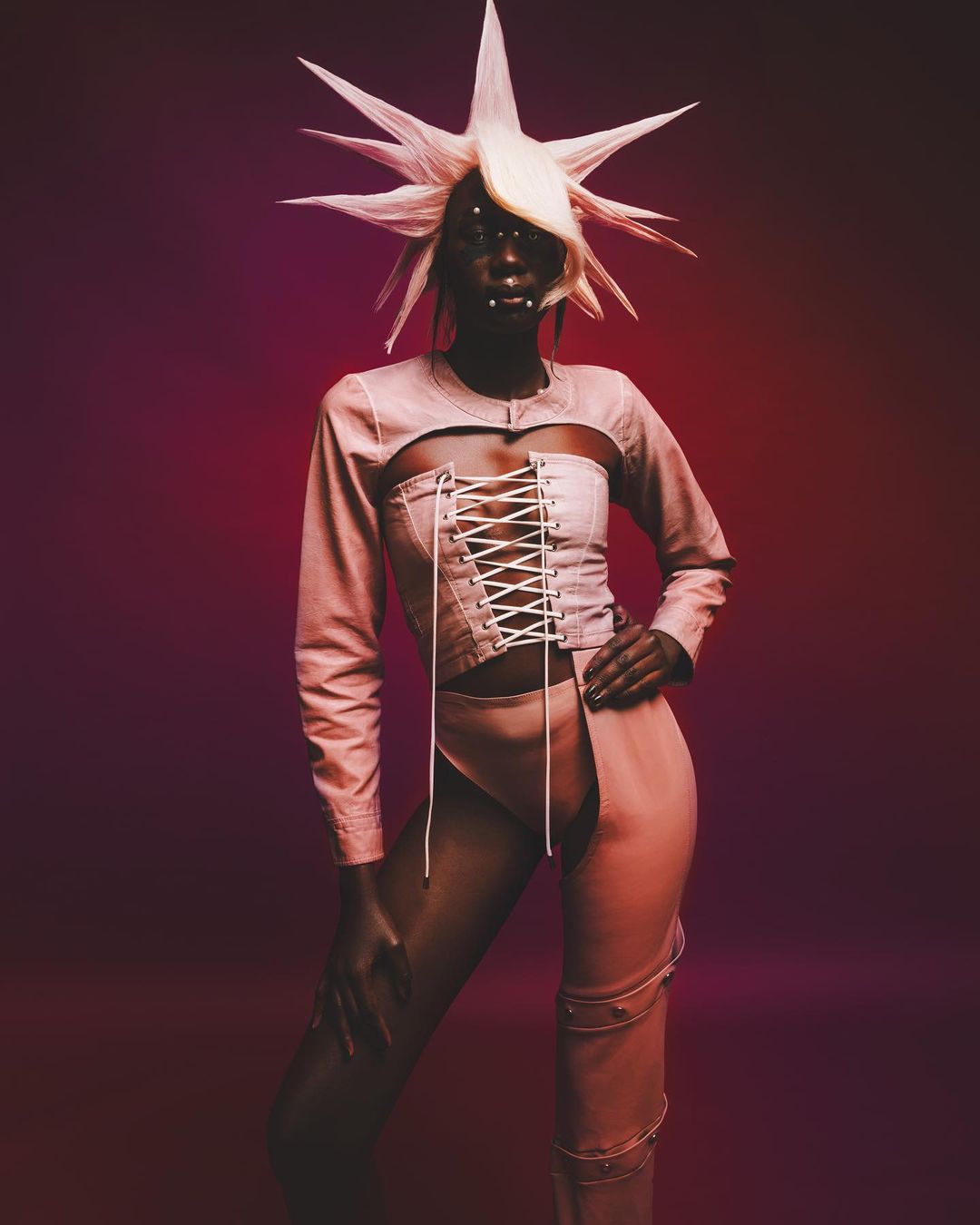 Unique fashion shot of an African woman in an avant-garde blonde wig with a pale pink two-piece outfit, posing in front of a burgundy backdrop.