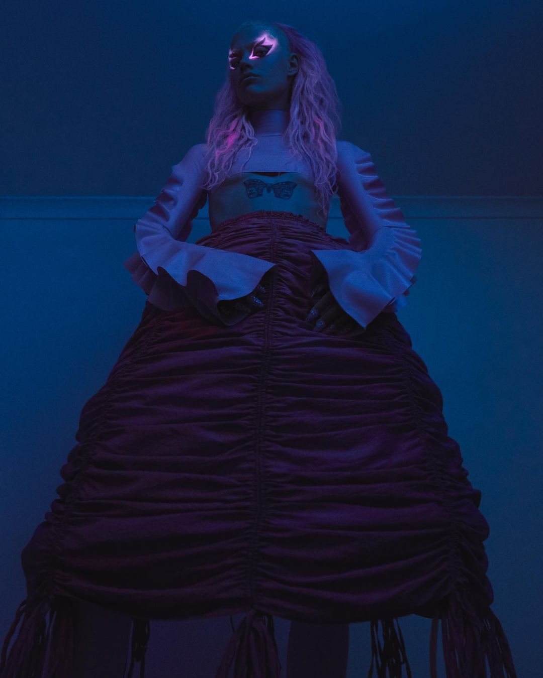 Woman in a ruched skirt and white bell-sleeve crop top stands in a low-angle shot in blue lighting with eye makeup and long hair.