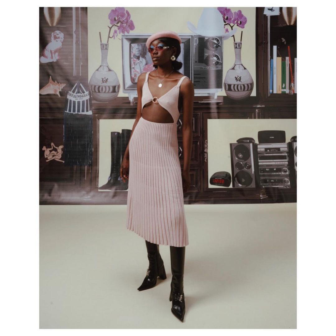 Woman in pink dress and black boots stands in front of a backdrop featuring furniture and electronics, posing for a fashion shot.