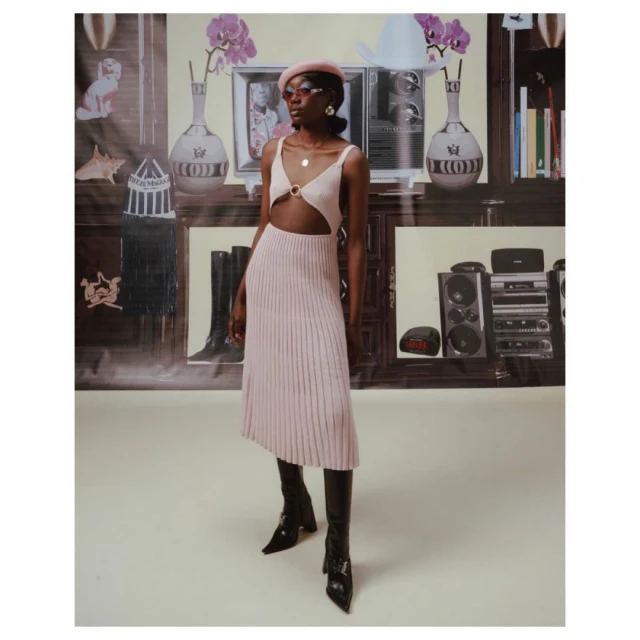 Woman in pink dress and black boots stands in front of a backdrop featuring furniture and electronics, posing for a fashion shot.