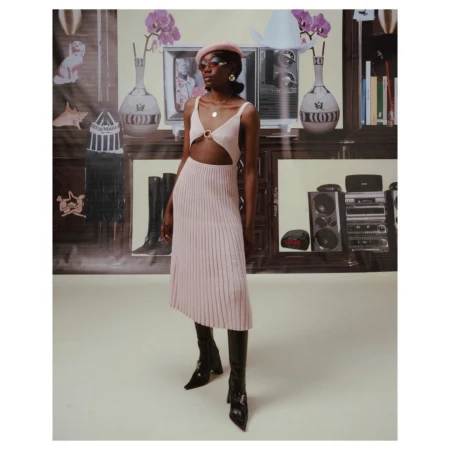 Woman in pink dress and black boots stands in front of a backdrop featuring furniture and electronics, posing for a fashion shot.