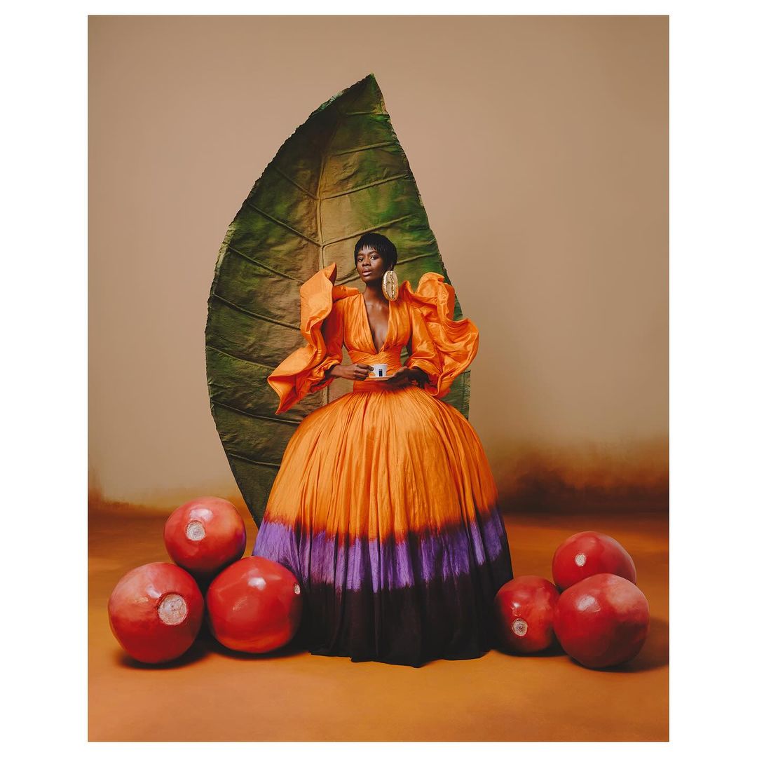 Woman in voluminous orange gown poses in studio with giant green leaf and bright red orbs, looking sophisticated and fashionable.