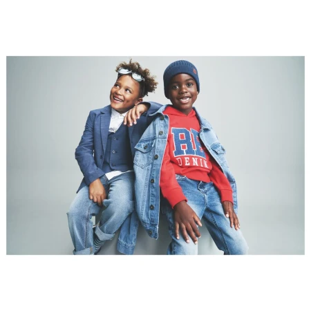 Two young boys smiling against a grey studio backdrop, one in a blue blazer and vest, the other in a red hoodie and denim jacket.