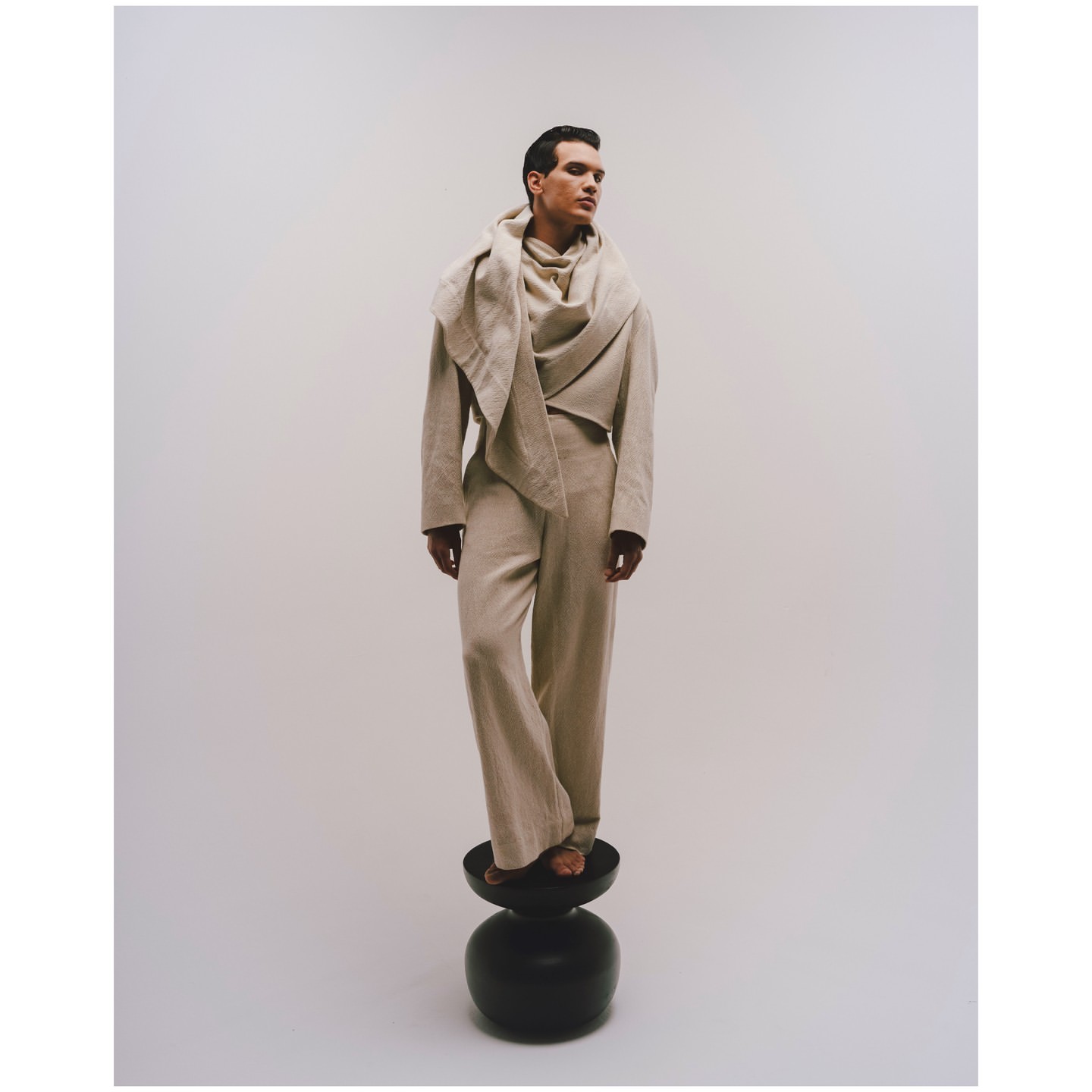 Fashion shot of a dark-haired adult male model standing on a black stool in a beige linen suit and draped scarf, against a white background.