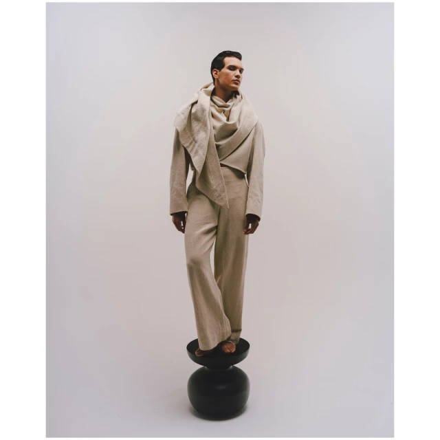 Fashion shot of a dark-haired adult male model standing on a black stool in a beige linen suit and draped scarf, against a white background.