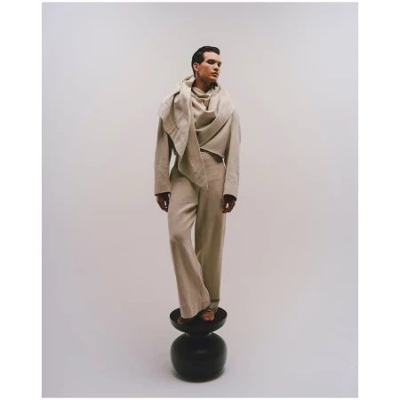 Fashion shot of a dark-haired adult male model standing on a black stool in a beige linen suit and draped scarf, against a white background.