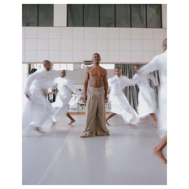 Athletic black man stands barefoot in wide-leg trousers surrounded by blurred figures in flowing white robes in dance studio.