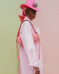Black woman wearing a pink cowboy hat and textured pink coat against a pastel wall