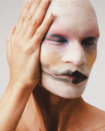 Close-up portrait of a bald person with avant-garde colourful makeup and a hand touching their head against a plain background